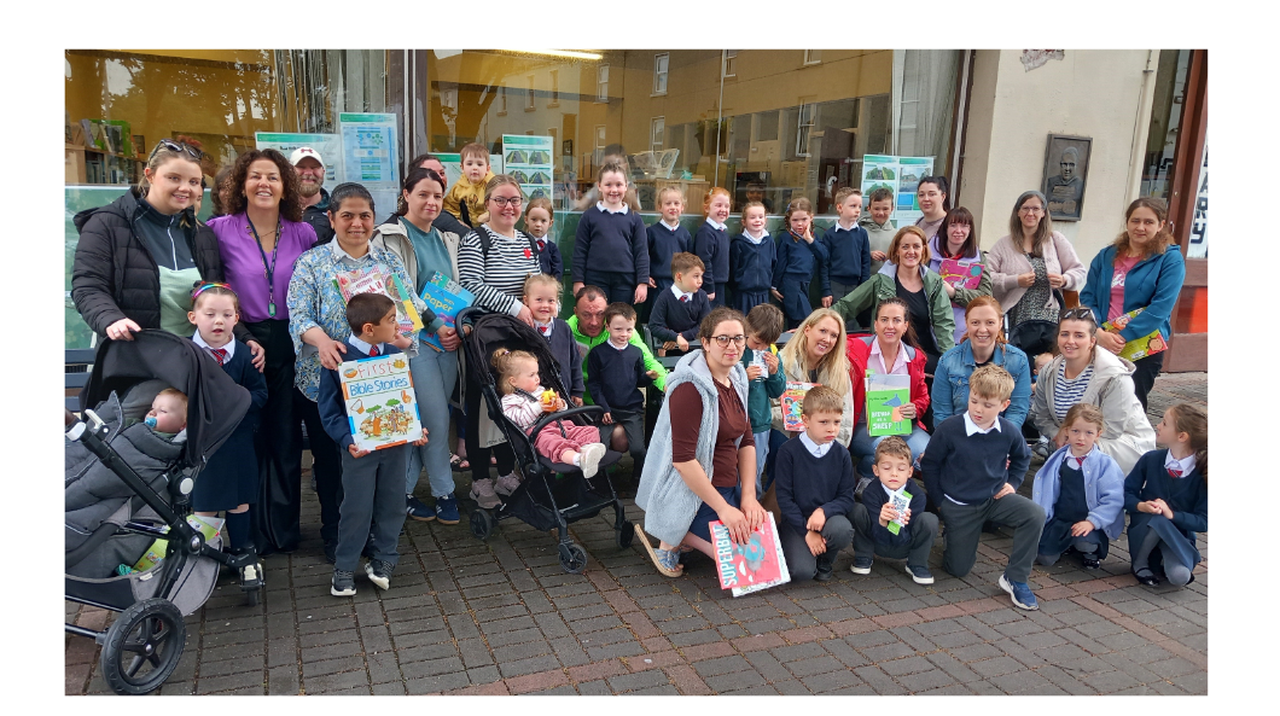 A group of adults and children gather outside a store, holding colorful children's books. Some children are in strollers, and others are standing.